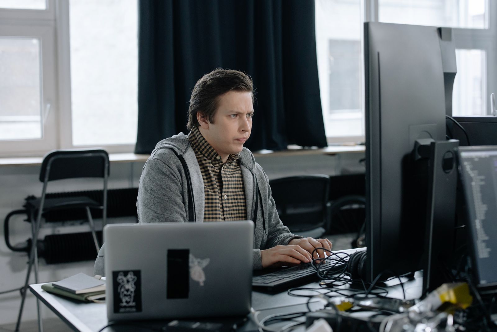 Homem de moletom cinza trabalhando em uma mesa com monitor e um notebook com adesivos, em um escritório aberto.