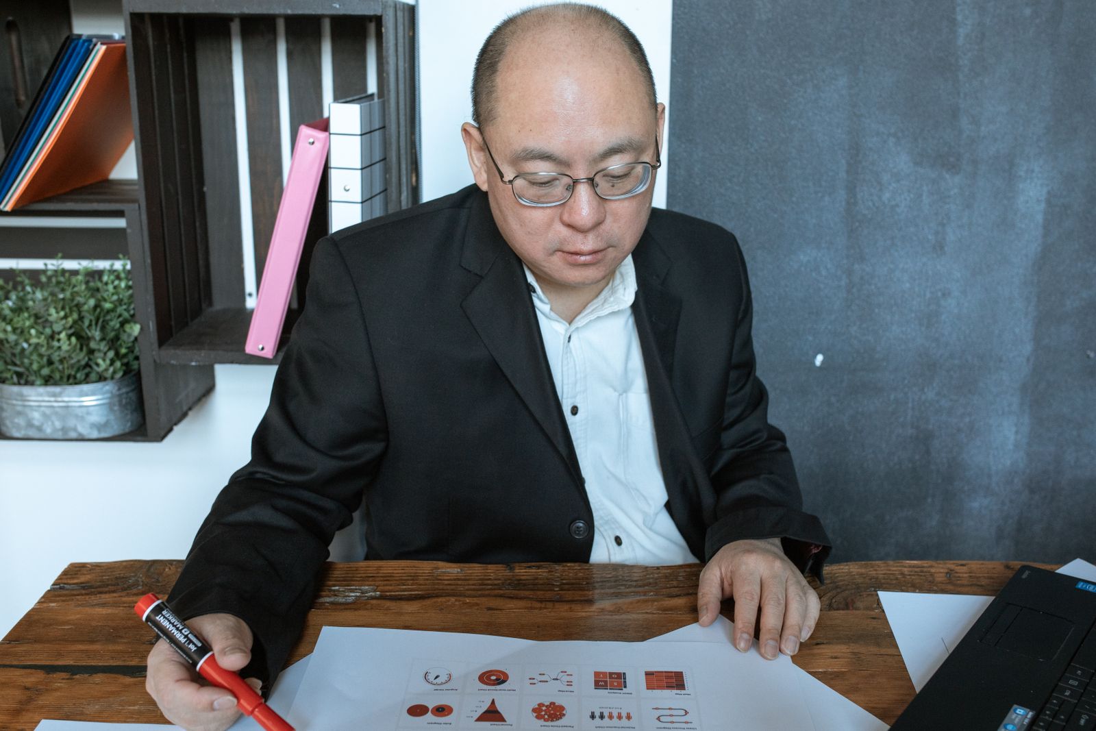 Homem de blazer escuro e óculos sentado a uma mesa de madeira, segurando um marcador vermelho sobre diagramas impressos, com um notebook no canto.