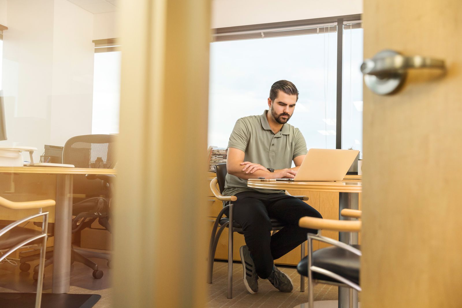 Homem de camisa polo trabalhando em um notebook em uma mesa, em um escritório visto por uma porta.