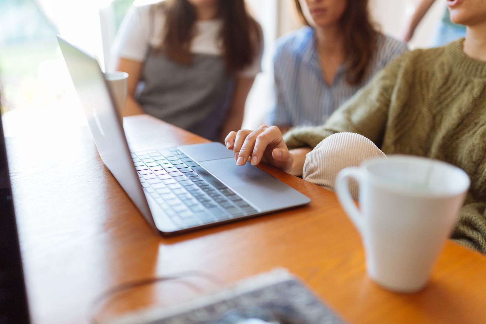 Mão sobre o touchpad de um notebook em uma mesa de madeira, com três pessoas sentadas ao fundo e uma caneca branca em primeiro plano.