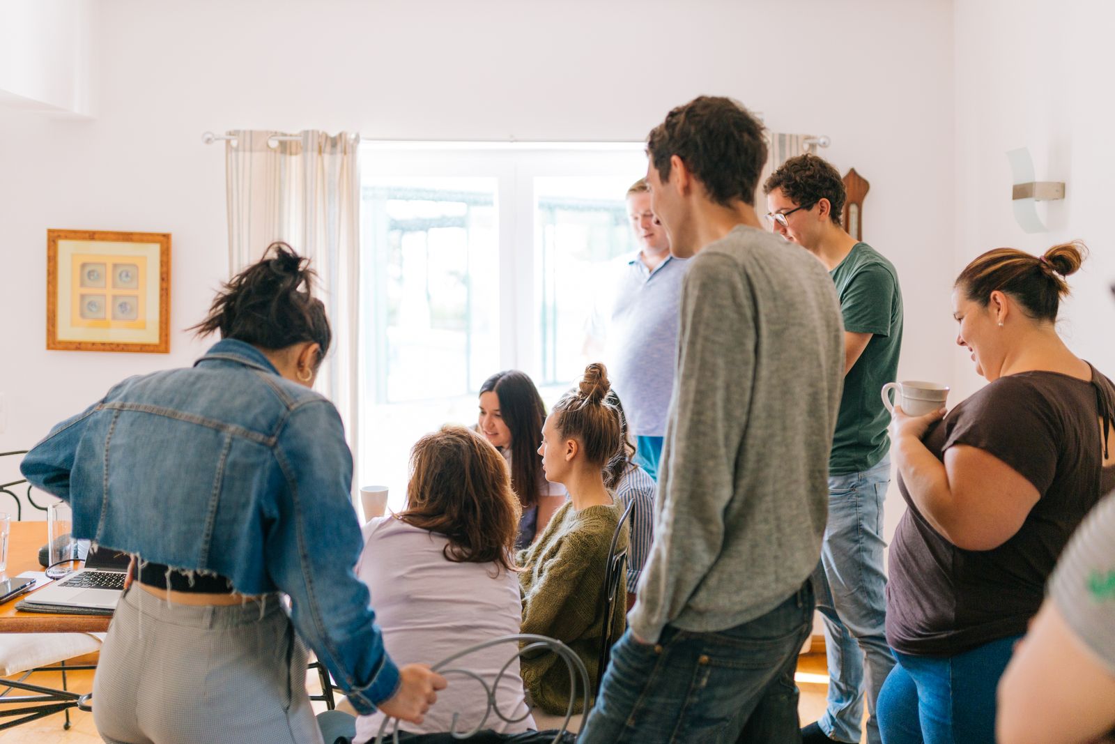 Grupo de cerca de oito pessoas em pé e sentadas em volta de uma mesa com um notebook, em uma sala clara com aspecto residencial, algumas segurando canecas.